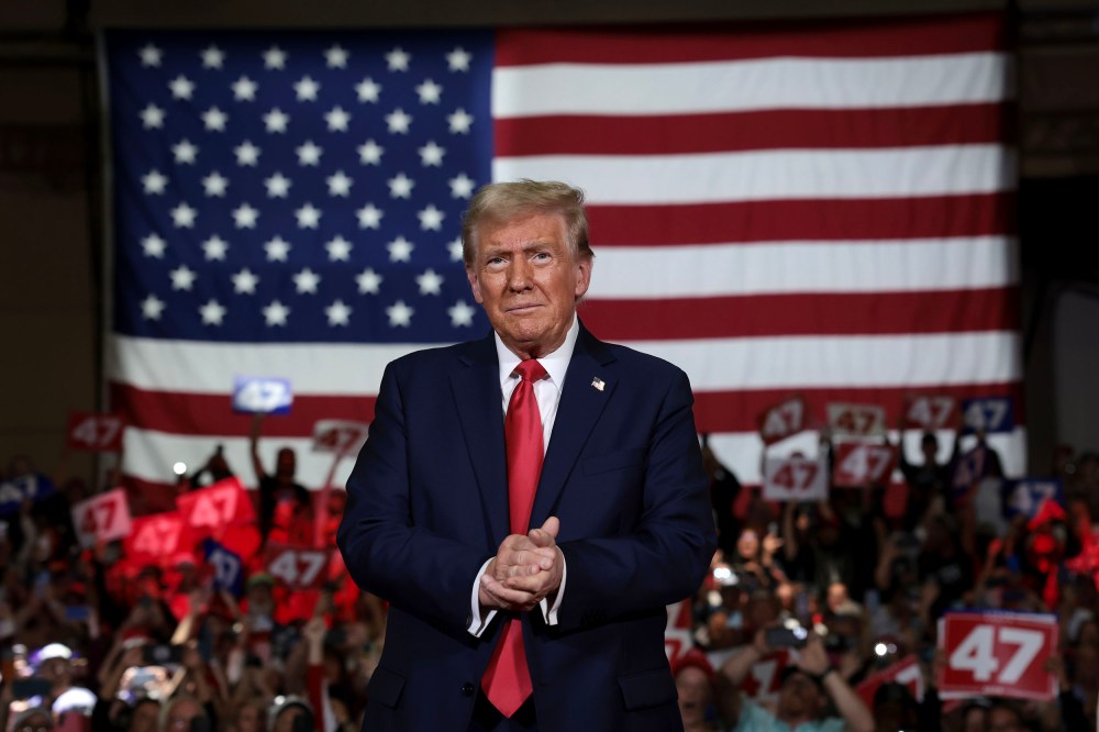 Donald Trump claps with an audience holding "47" signs behind him. A large American flag is the backdrop.