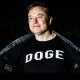 Elon Musk smiles and opens up his jacket to show a black shirt that reads "DOGE" in white lettering
