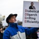A person holds a sign outside that reads "Diabolical Oligarch Grifting Efficiently"