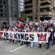 A large group of people walk through a street and hold a banner that says "No Kings"