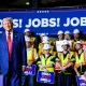 Donald Trump stands on stage in front of workers wearing safety vests