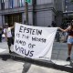 Two people hold a white blanket sign with black text that reads "Epstein is the worst kind of virus"