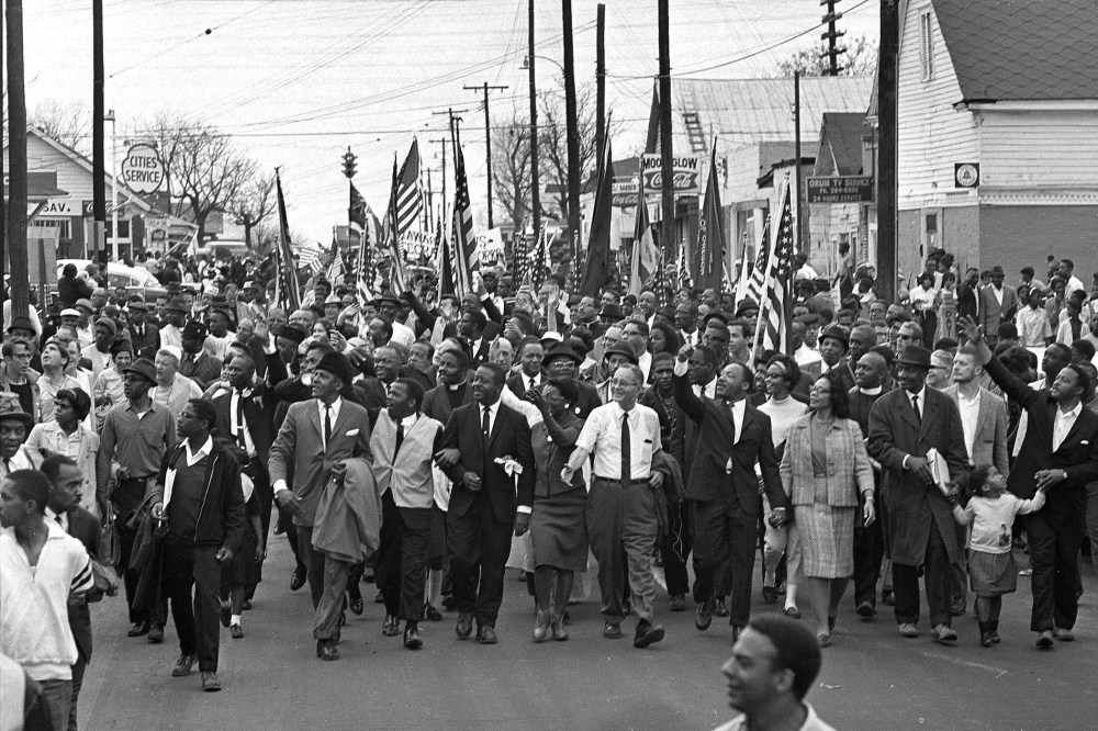 In this March 21, 1965 file photo, Dr. Martin Luther King, foreground row, fifth from right, waves as marchers stream across the Alabama River on the first of a five day, 50-mile march to the state capitol at Montgomery, Ala. (Photo by AP)