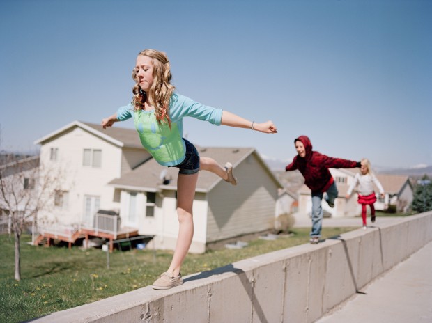 A young teeanage girl does gymnastics moves while balancing on a concrete ledge as her little brother and sister copy her with a view of mountains and her family's suburban neighborhood in background. Pleasant Grove, Utah. Brian Shumway/Redux