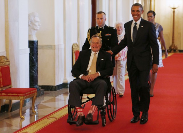 Former U.S. President George H.W. Bush sits in a wheelchair as he is escorted next to U.S. President Barack Obama (R) as they attend an event honoring the 5,000th winner of the "Daily Point of Light" award in the East Room of the White House in...