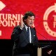Tucker Carlson speaks at a podium in front of a red Turning Point USA logo screen.