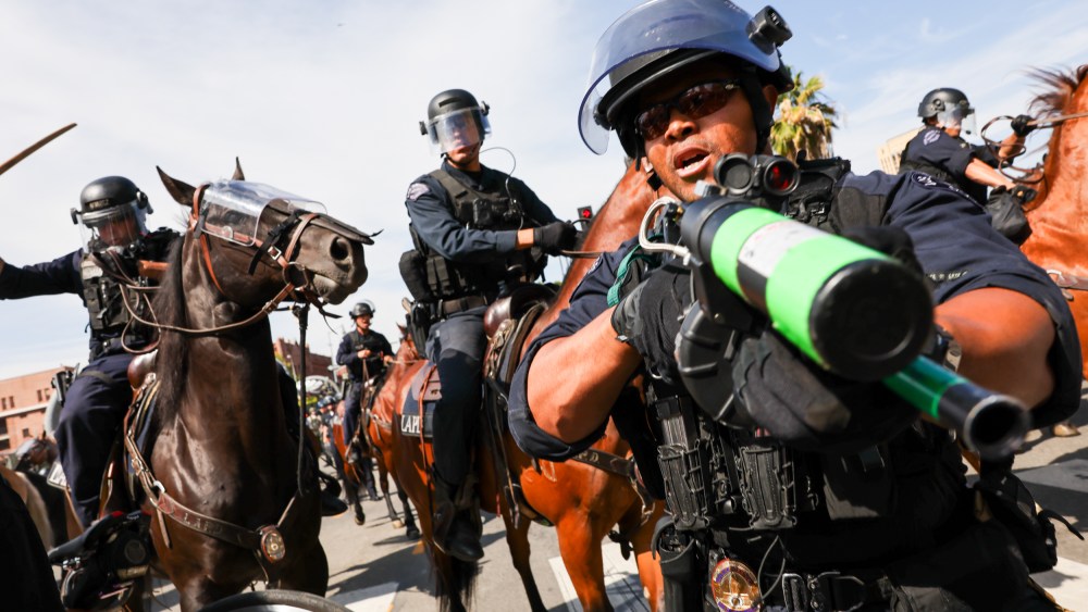 A police officer holds a weapon to the camera as protesters face off with police outside of a federal building in downtown Los Angeles for an anti-Trump "No Kings Day" demonstration.