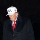 Donald Trump walks at the White House at night, wearing a white USA hat.