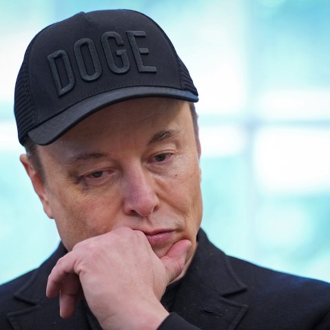 Elon Musk, wearing a black hat that says "DOGE," puts his hand to his chin while looking down.