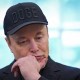 Elon Musk, wearing a black hat that says "DOGE," puts his hand to his chin while looking down.