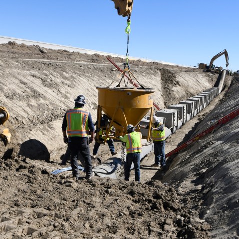 Workers in yellow high-visibility safety garments and hard hats set a concrete bucket on the ground of a construction zone.