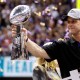 John Harbaugh holds the Vince Lombardi Championship trophy as purple and gold confetti falls in the background.
