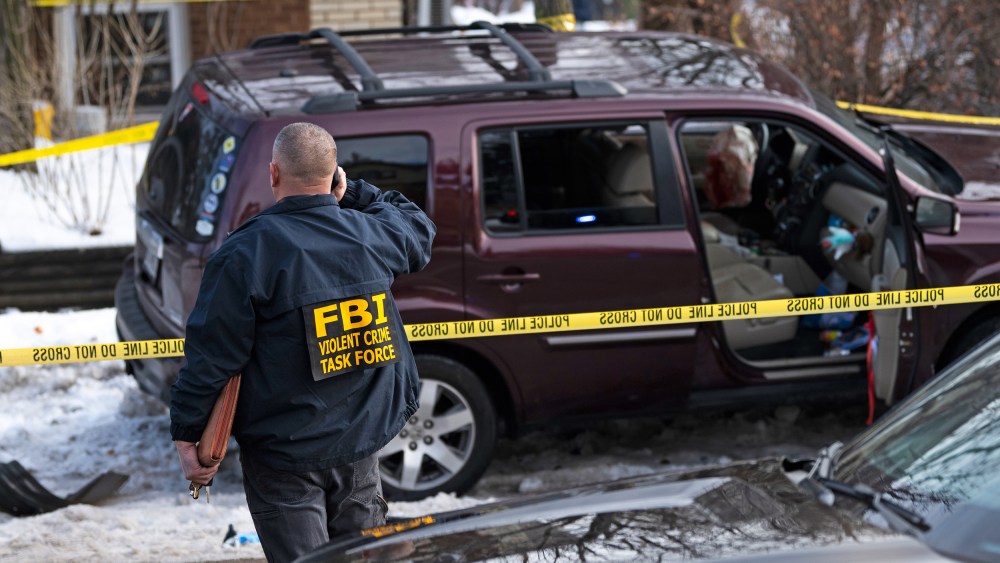 An FBI agent stands near a wrecked car surrounded by yellow police line tape on the side of the road.