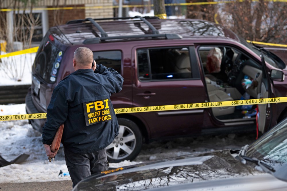 An FBI agent stands near a wrecked car surrounded by yellow police line tape on the side of the road.