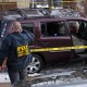 An FBI agent stands near a wrecked car surrounded by yellow police line tape on the side of the road.