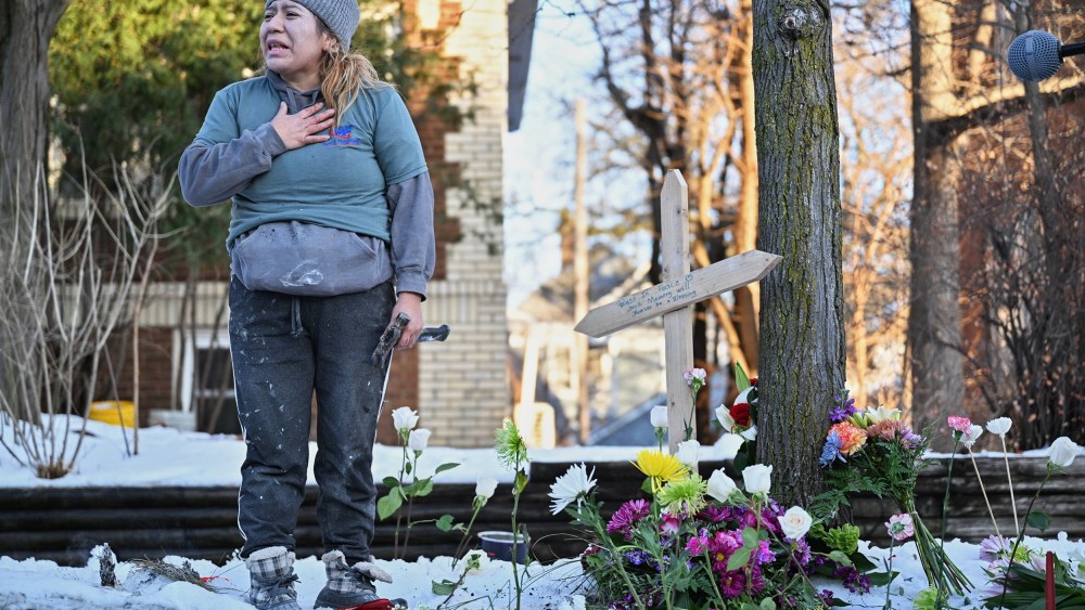 A woman in distress with her hand on her heart stands next to a wooden cross surrounded by flowers.