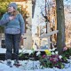 A woman in distress with her hand on her heart stands next to a wooden cross surrounded by flowers.