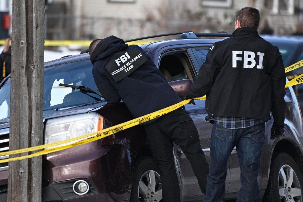 People wearing FBI clothing lean over caution tape onto a car on the side of the road.
