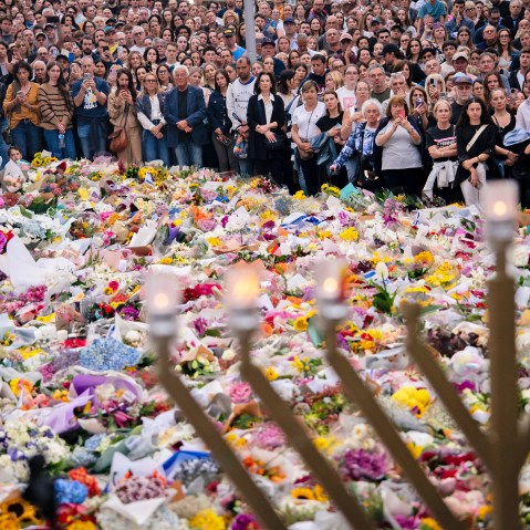 A crowd of people surrounds a large memorial filled with flowers. A menorah stands in the foreground.
