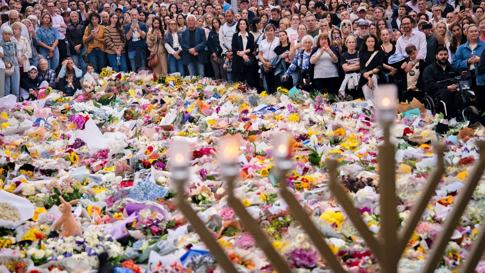 A crowd of people surrounds a large memorial filled with flowers. A menorah stands in the foreground.