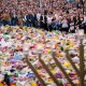 A crowd of people surrounds a large memorial filled with flowers. A menorah stands in the foreground.