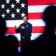 Silhouettes of people watch Elon Musk holding a microphone on a stage backdropped by a digital American flag.