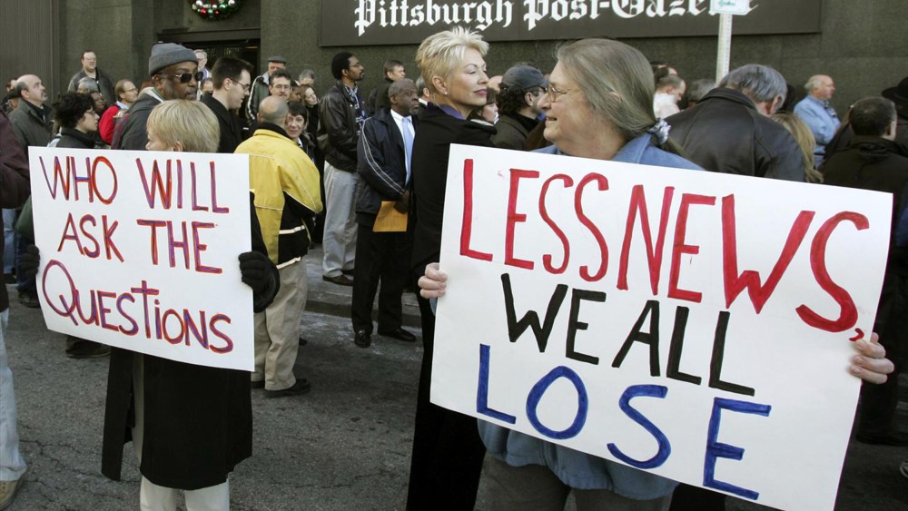 People hold signs reading "Who will ask the questions" and "Less news we all lose" outside the Pittsburgh Post-Gazette building.