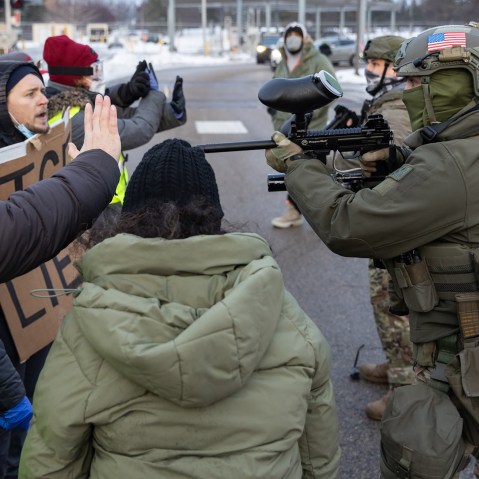 A federal agent points a weapon towards protestors who raise their hands to say "stop."