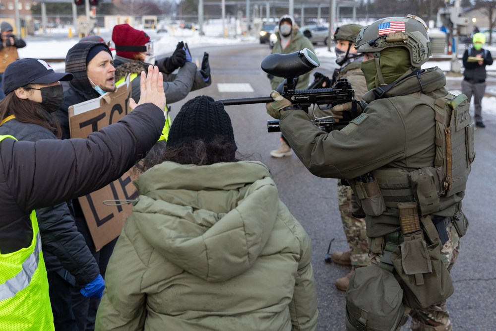 A federal agent points a weapon towards protestors who raise their hands to say "stop."