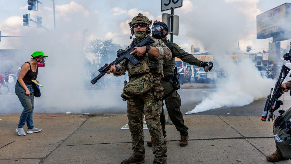 One federal agent stands while another one sprays a foreign agent. There is a protestor wearing a gas mask in the gas cloud.