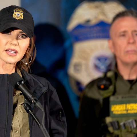 Kristi Noem talks at the podium while Gregory Bovino stands in the background in Minneapolis.