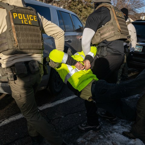 Federal agents carry a handcuffed person by the person's arms and legs.