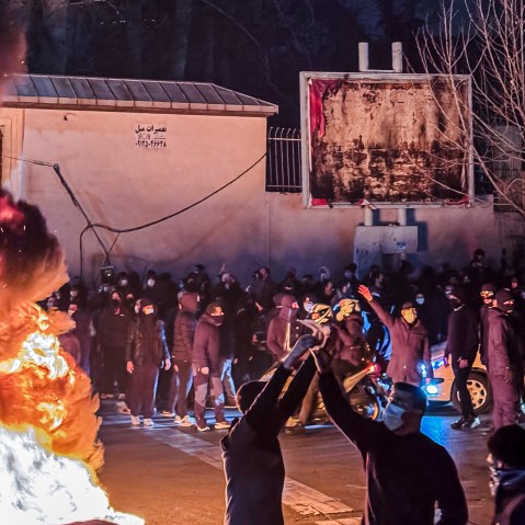 Two Protestors raise their hands in front of a fire. There are other on the sidelines.