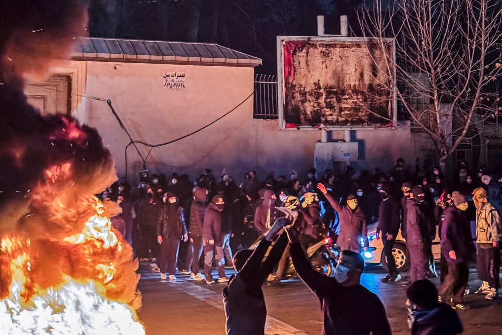 Two Protestors raise their hands in front of a fire. There are other on the sidelines.