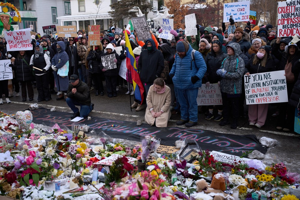 A group of protesters holding signs visit a memorial for Renee Nicole Good. Several of them are kneeling on the ground.