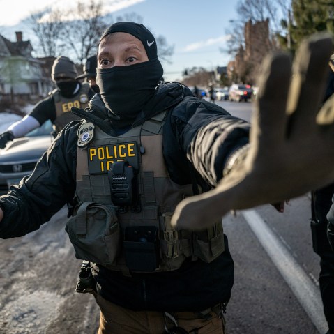An ICE agent signals "stop" with his left hand up while holding a spray bottle as other agents follow him.