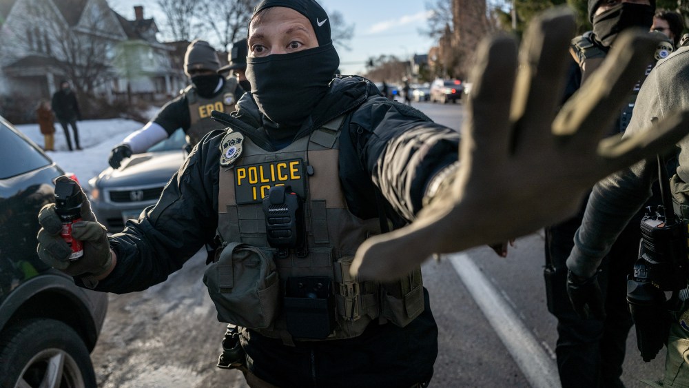 An ICE agent signals "stop" with his left hand up while holding a spray bottle as other agents follow him.