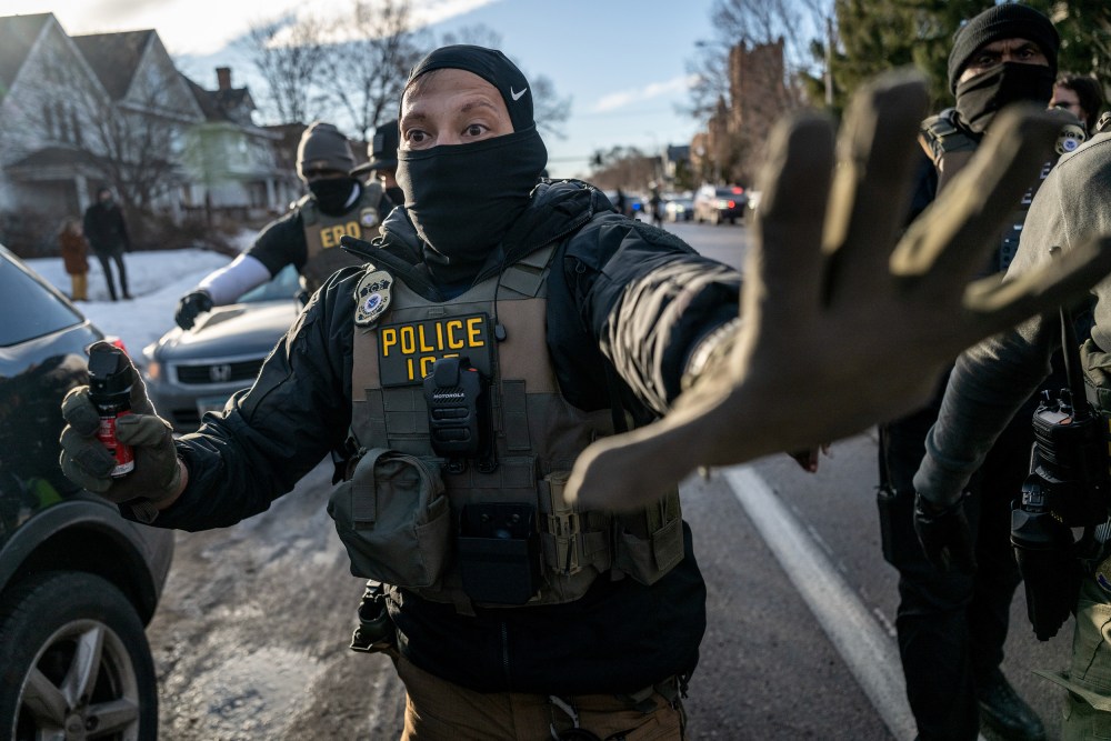 An ICE agent signals "stop" with his left hand up while holding a spray bottle as other agents follow him.