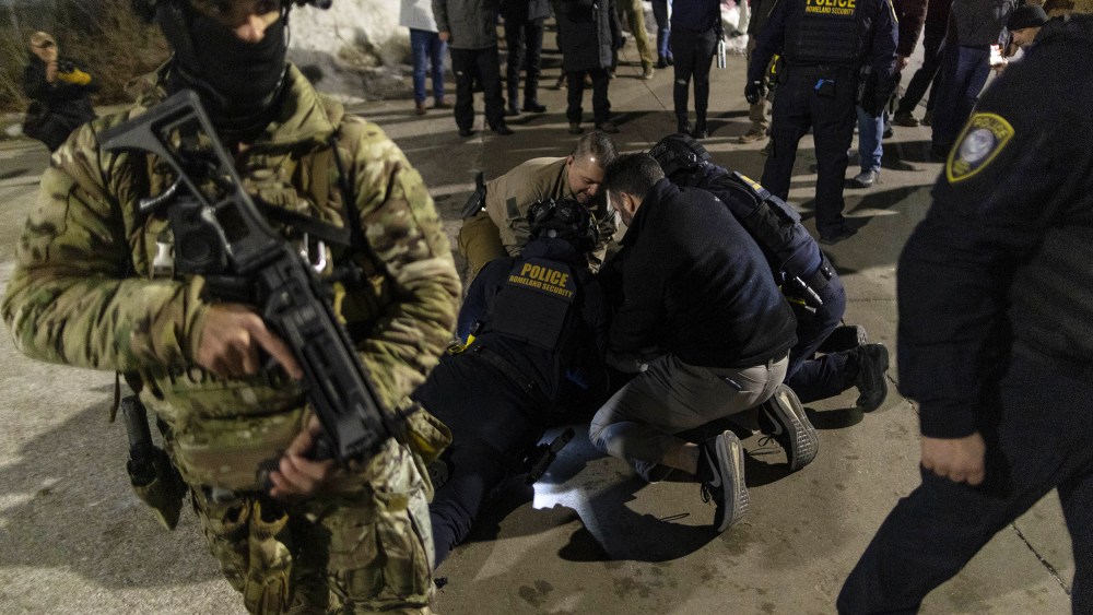 A federal agent holds a weapon in the foreground while multiple agents try to arrest a protester in the background.
