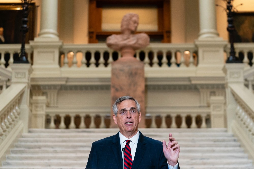 Brad Raffensperger speaks at the bottom of a staircase while gesturing with his hand.