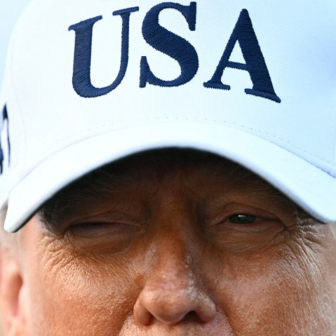 A closeup photo of President Trump shows the upper half of his face wearing a white USA hat.